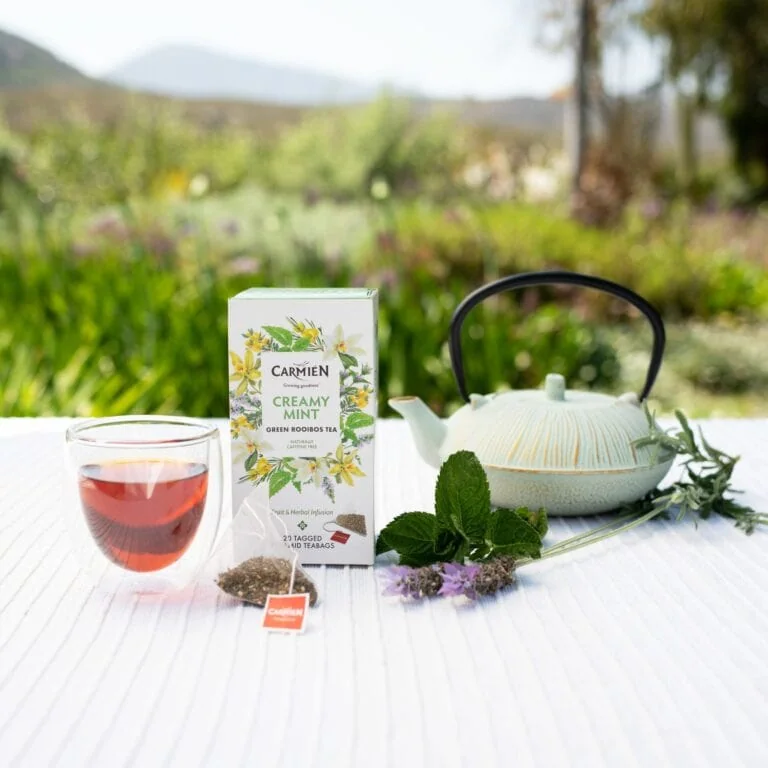CarmiƩn Tea Creamy Mint rooibos tea with fresh mint leaves and teapot on outdoor table, promoting herbal tea blends for health and relaxation.