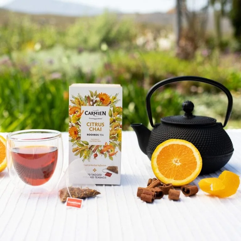 Carmién Citrus Chai Rooibos Tea with fresh orange, teapot, and tea bag on a white table outdoors.