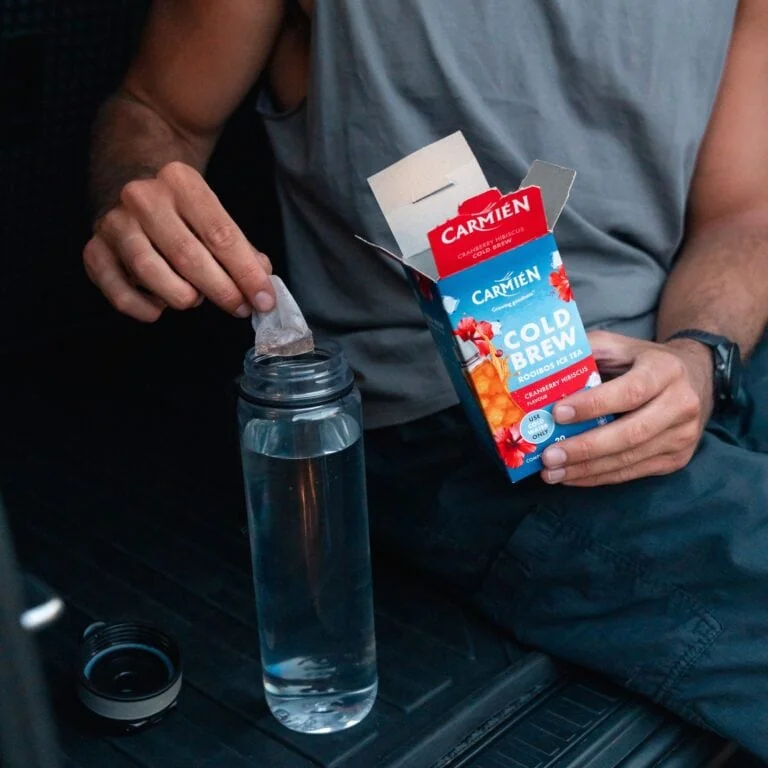 Person opening Carmién Cold Brew Rooibos Ice Tea box with a teabag in a water bottle, promoting healthy rooibos tea consumption.