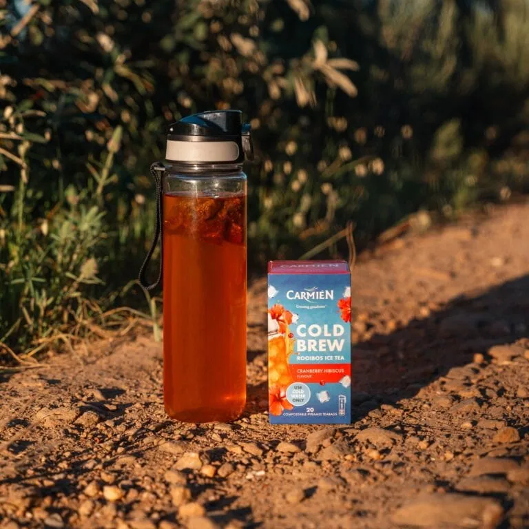 Carmién Rooibos Cold Brew Tea in a glass bottle with a box of Cranberry Hibiscus flavor on a dirt path.