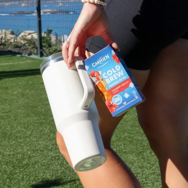 Carmién Cold Brew Rooibos Ice Tea box held outdoors with a white tumbler on green grass, showcasing a refreshing tea product for e-commerce.