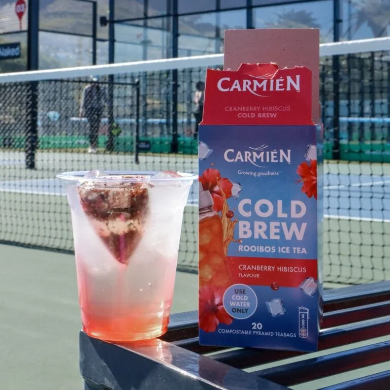 Carmién Cranberry Hibiscus Cold Brew Rooibos Ice Tea box with a glass of iced tea on a tennis court.