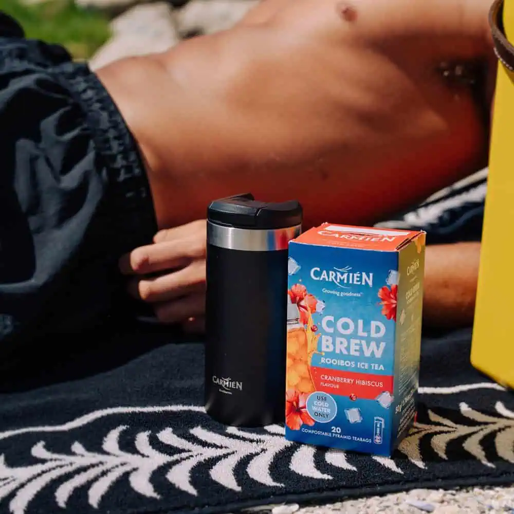 An image of a Carmién Cranberry Hibiscus Cold Brew Rooibos Tea box next to a black travel mug, set outdoors on a sunny day.
