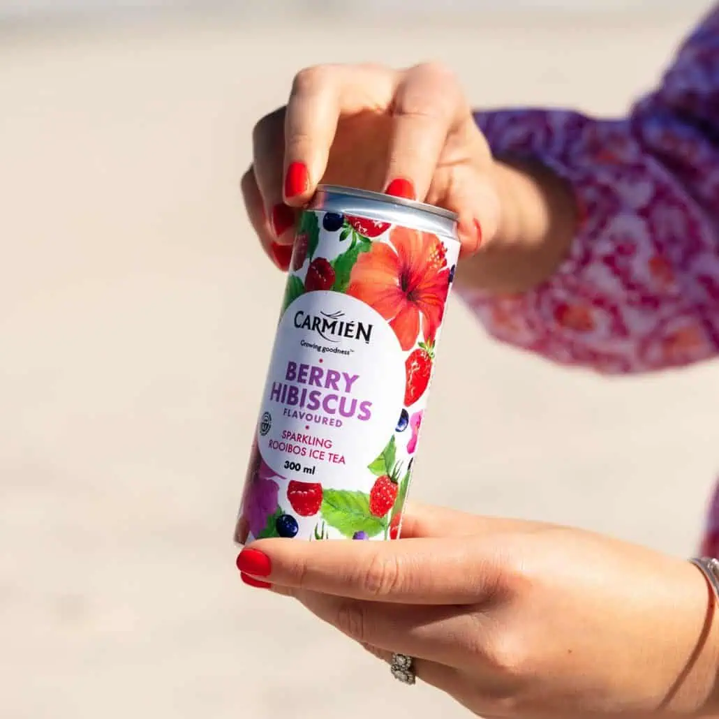 Berry Hibiscus flavoured Carmién Tea sparkling rooibos ice tea in a 300ml can, held by a hand with bright red nail polish, against a neutral background.