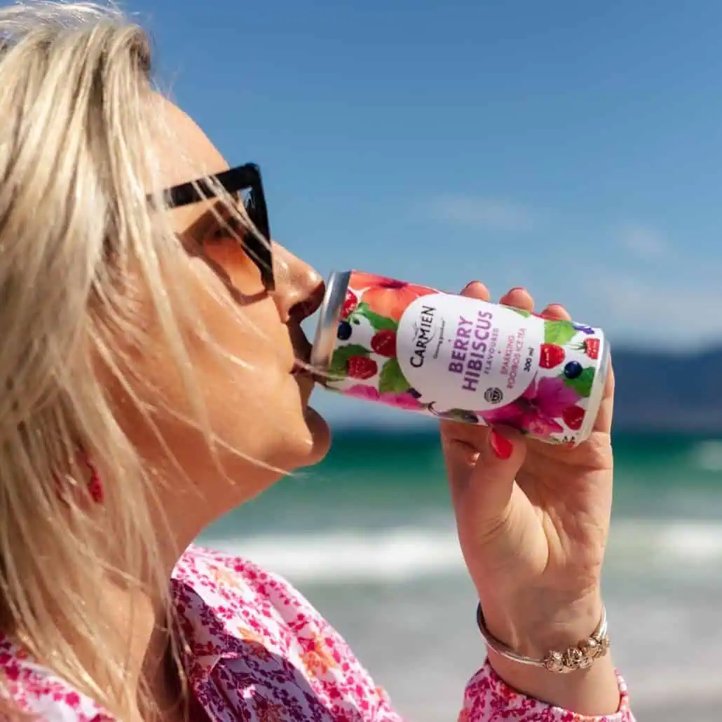 Carmién Berry Hibiscus sparkling tea being enjoyed outdoors at the beach with blue sky and ocean in the background.