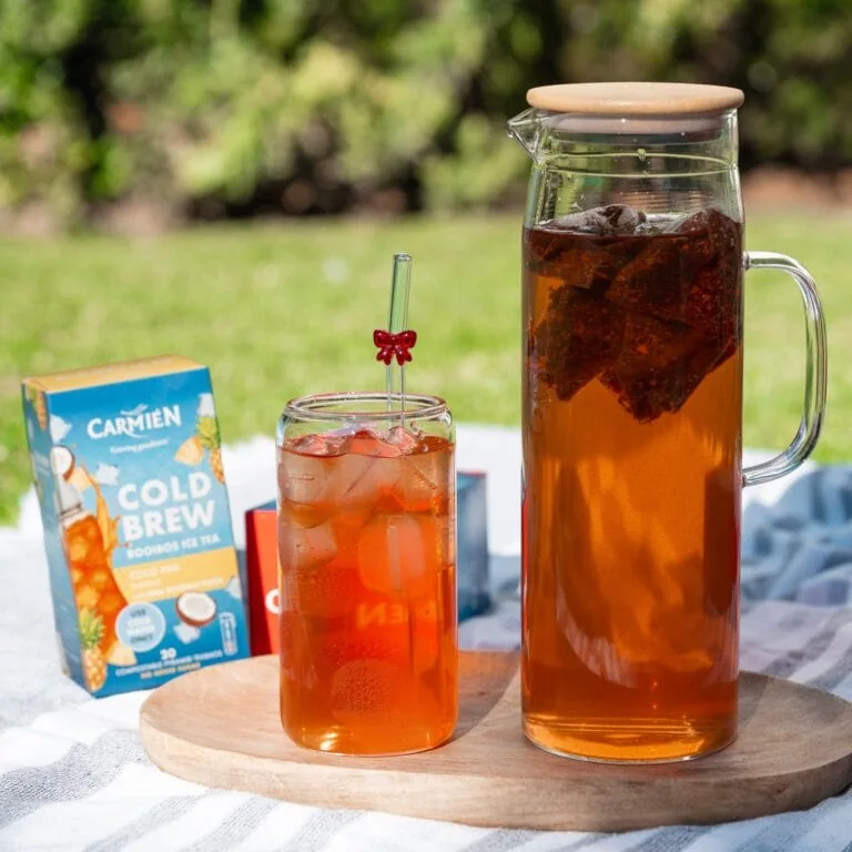 Carmién Rooibos tea served in a glass jug and a glass with ice, outdoors on a sunny day.