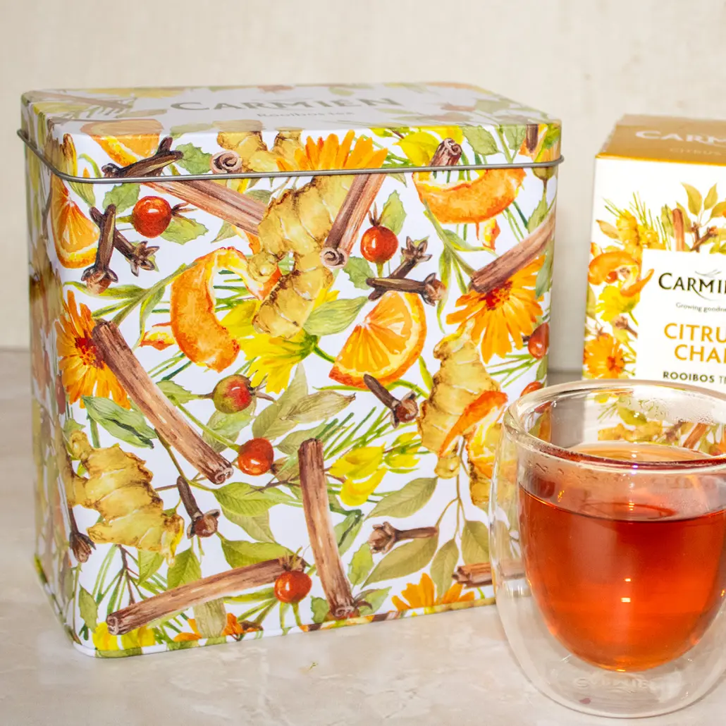 Fresh Carmién Tea citrus-flavored rooibos tea in a decorative box with a glass of brewed tea on a neutral background.