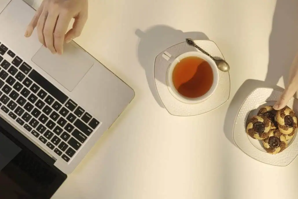 A person enjoying a cup of CarmiƩn Rooibos Tea alongside a plate of cookies and a laptop, highlighting a healthy alternative to coffee for avoiding the 3pm crash.