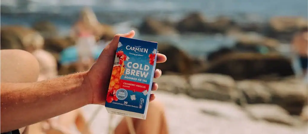 Hand holding a Carmién Cranberry Hibiscus Cold Brew Pack on a beach, showcasing the refreshing tea with cranberry and hibiscus flavors.
