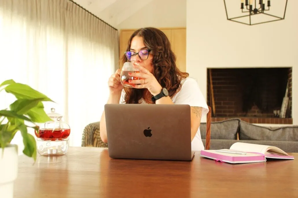 A woman enjoying a cup of Carmién Tea while working on her laptop at home, with a cozy living room background.