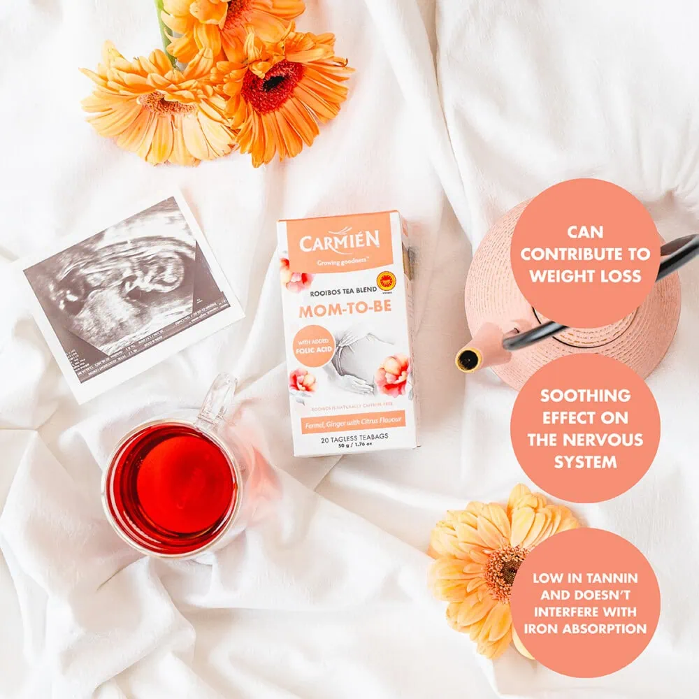 Carmién Rooibos Mom-to-Be tea box with flowers, ultrasound image, and tea accessories on a white fabric background.