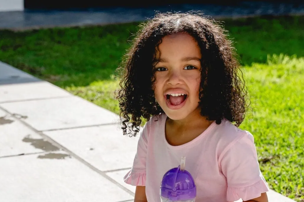 Young girl with curly hair smiling outdoors, holding a purple water bottle on a sunny day.