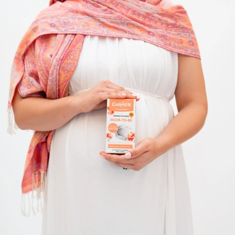 Pregnant woman holding Carmién Mom-to-Be tea box, promoting rooibos tea for pregnancy and nursing health benefits.