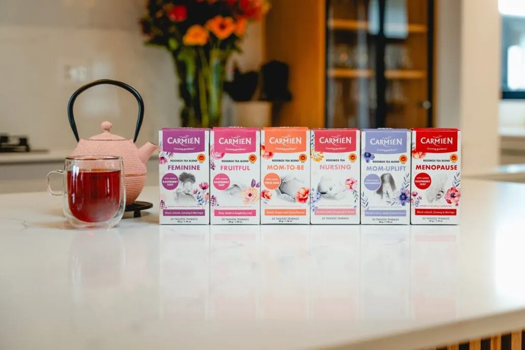 Assorted Carmién Rooibos tea boxes for women, including Feminine, Fruitful, Mom-to-Be, Nursing, Mom-Uplift, and Menopause blends, displayed on a kitchen counter.