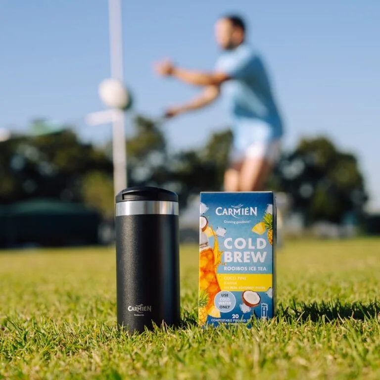 Carmién Cold Brew Rooibos Ice Tea box and travel mug on grass with a person playing volleyball in the background.