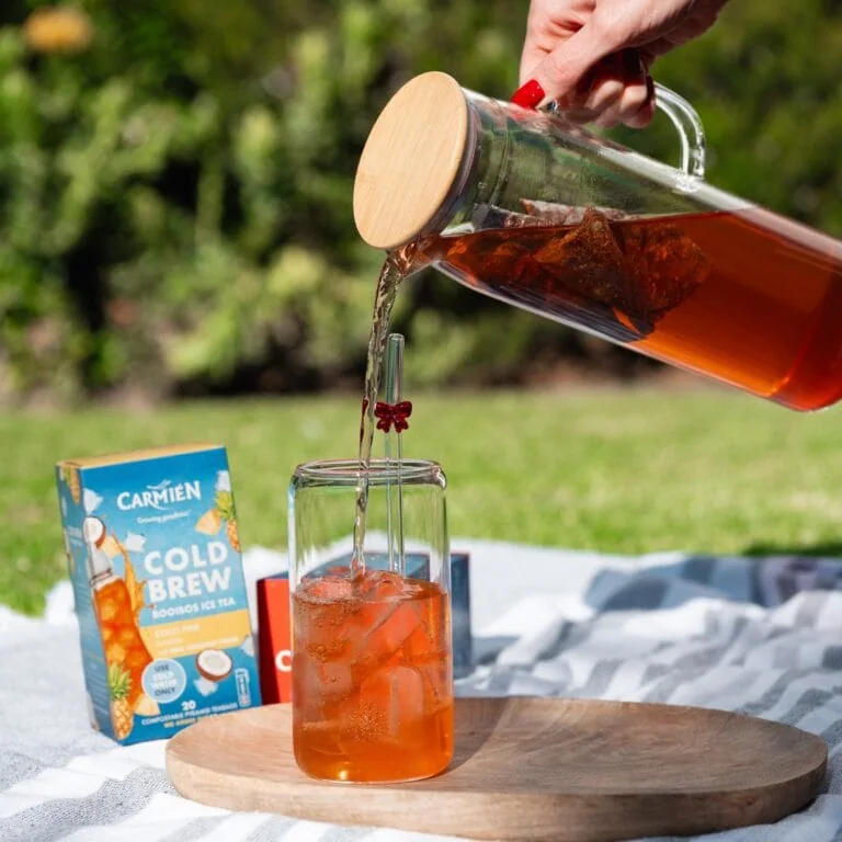 Refreshing CarmiƩn Rooibos Cold Brew Tea being poured over ice in a glass, with packaging visible in the background, perfect for summer refreshment.