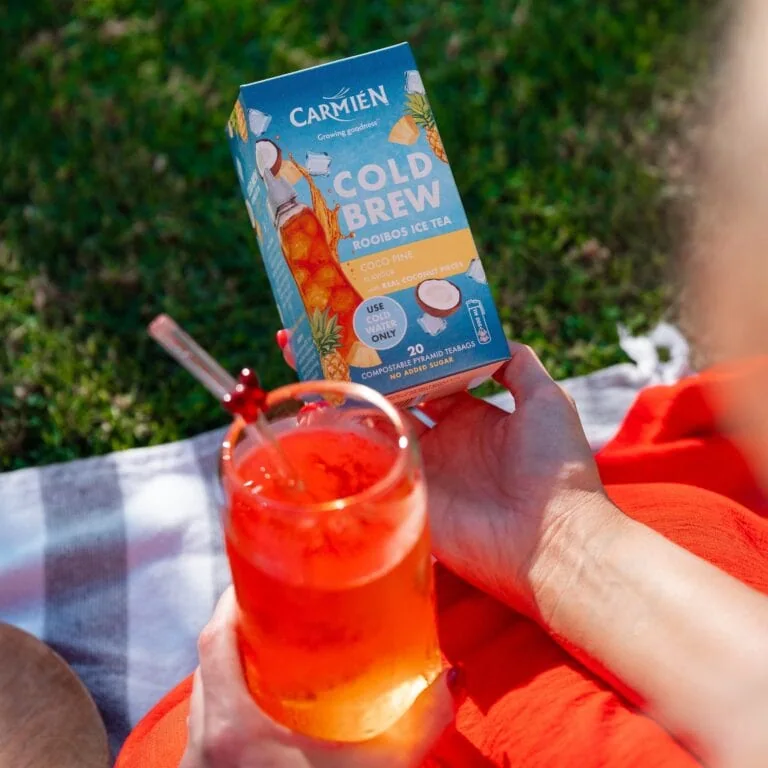 CarmiƩn Cold Brew Rooibos Ice Tea box held by a person outdoors with a glass of iced tea in the foreground.