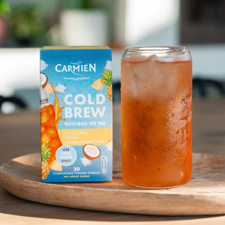 CarmiƩn Cold Brew Rooibos Ice Tea with coconut pineapple flavor in a glass, next to the tea box on a wooden surface.