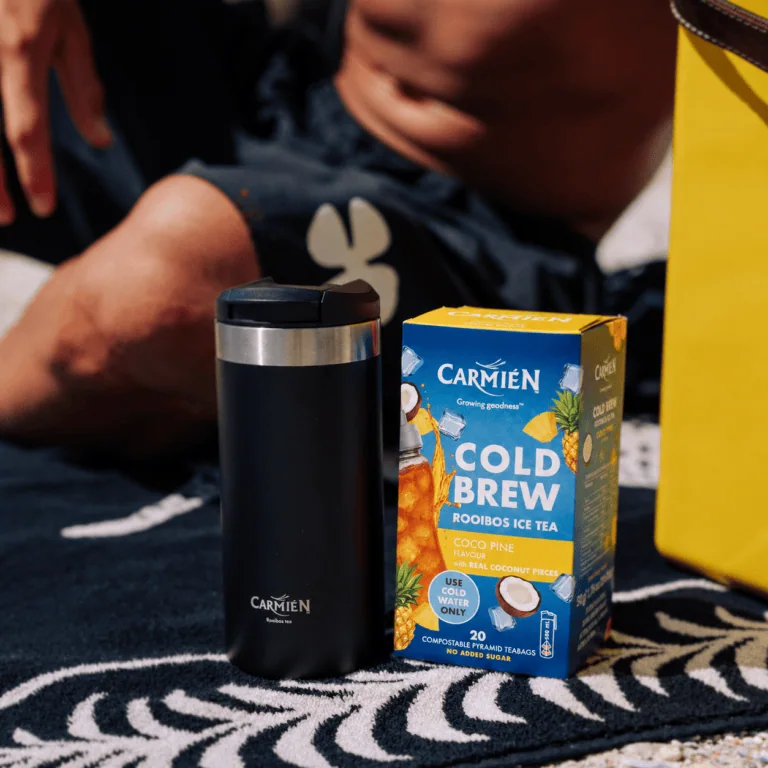 CarmiƩn Cold Brew Rooibos Ice Tea with Coco Pine flavor in a black insulated bottle and packaging on a patterned surface.