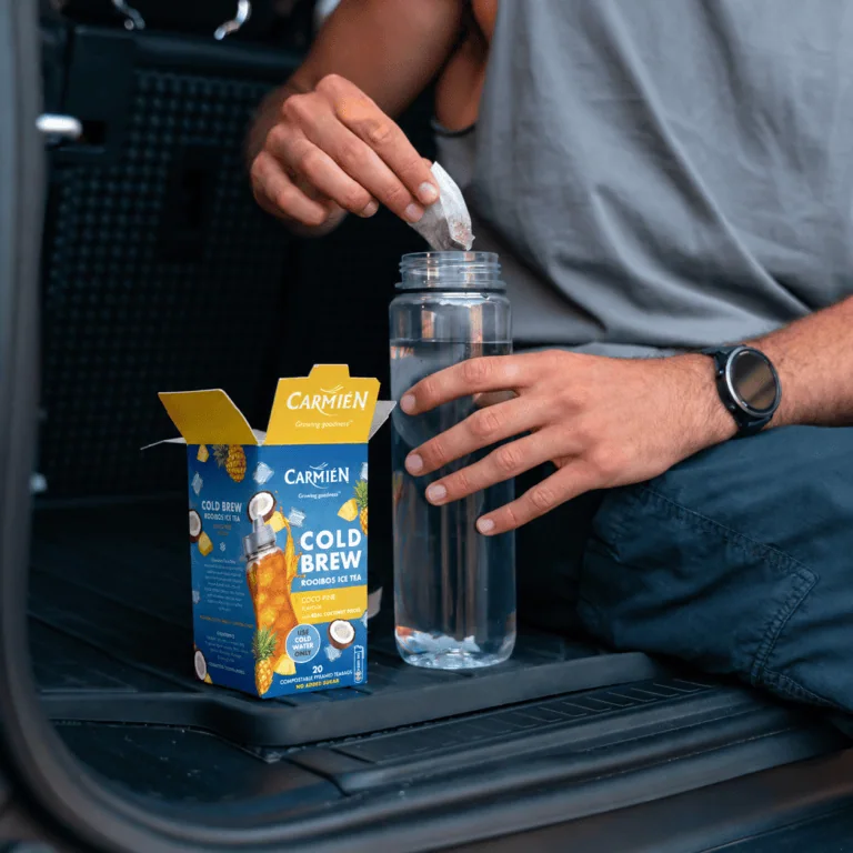 Person adding a tea bag to a glass water bottle with CarmiƩn Tea Cold Brew Rooibos Ice Tea packaging nearby.